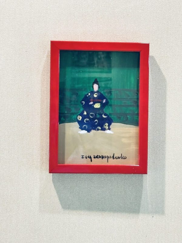 赤い額に入った絵画。緑の背景に、紺の装束と黒い烏帽子の行司が座る。Zoia Skoropadenkoの署名入り