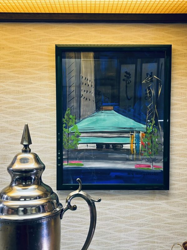 緑の屋根の両国国技館と背後の高層ビルを描いた額装の絵画。手前に銀のトロフィー
