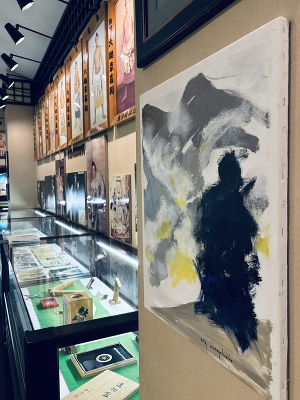 展示ケースと肖像が並ぶ大鵬相撲記念館の展示室。手前に黒・灰・黄で描かれた大きな人物画