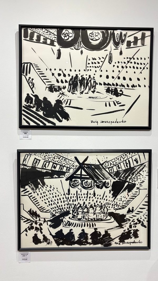 上下に掛けられた二点の額装墨画。土俵、吊り屋根、観客で埋まる相撲場の内部